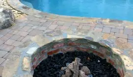 Stone fire pit filled with lava rock on a paver pool deck