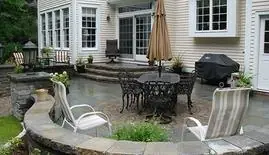 Stone patio with dining set, umbrella and grill behind a white home
